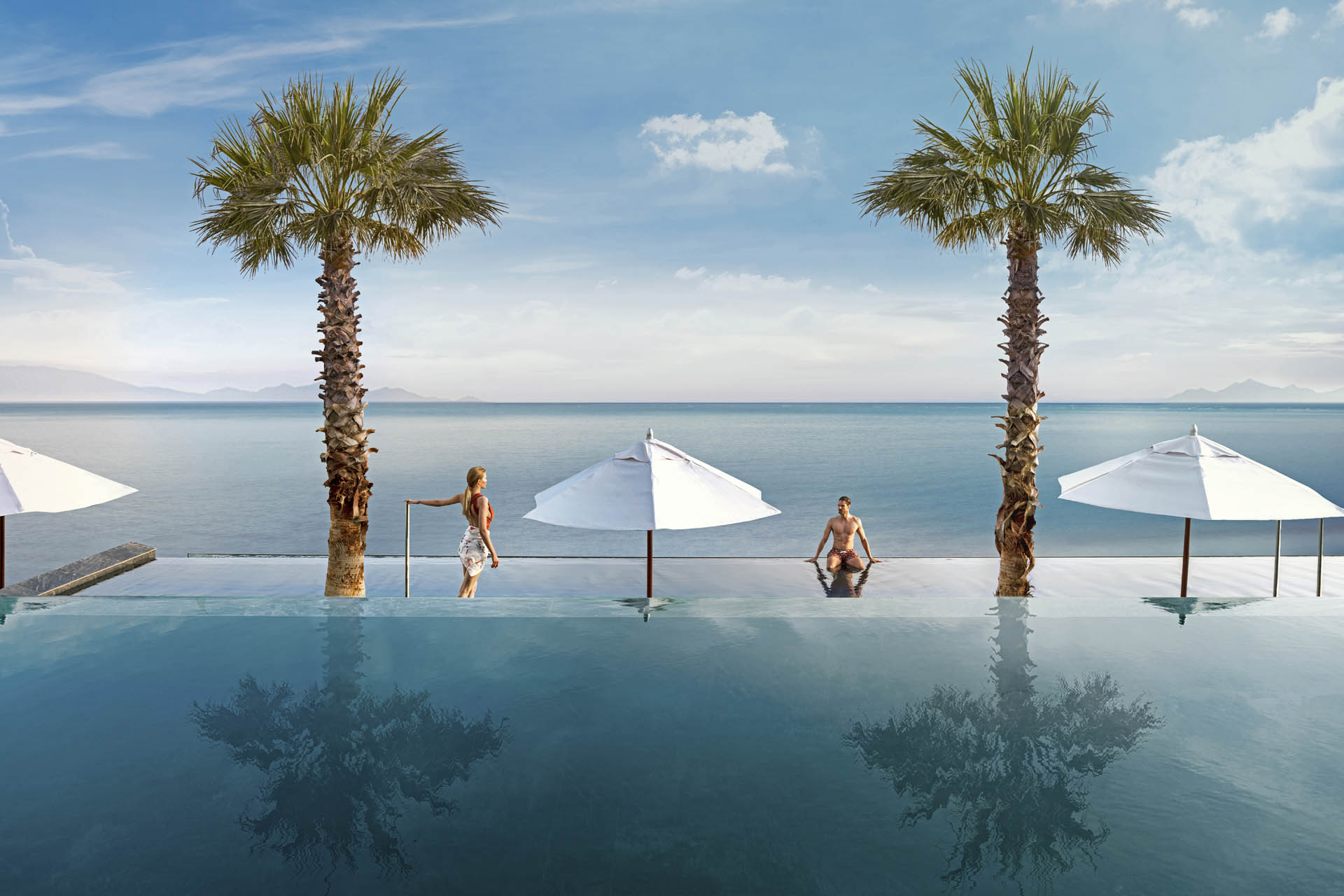 Poolanlage mit Infinity Pool mit weißen Sonnenschirmen und einem jungen Pärchen in einem Mitsis Hotel.